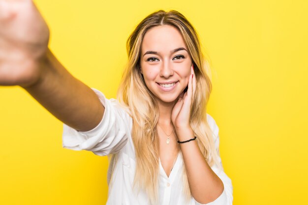 Portret van een glimlachende jonge vrouw die een selfie over gele muur neemt