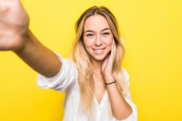 Portret van een glimlachende jonge vrouw die een selfie over gele muur neemt