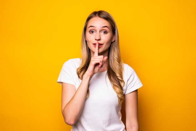 Portret van een glimlachende jonge vrouw die stiltegebaar toont dat over gele muur wordt geïsoleerd