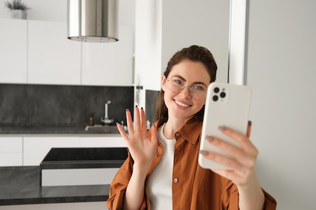 Gratis foto portret van een glimlachende jonge vrouw die thuis met een mobiele telefoon staat en met haar hand op een smartphone zwaait