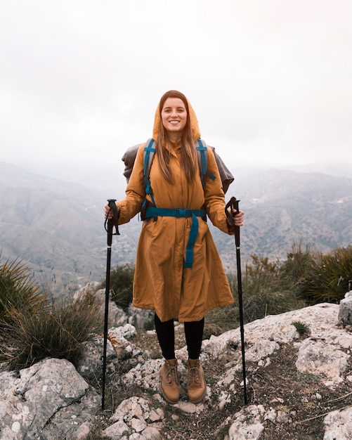 Portret van een glimlachende jonge vrouw met de wandelingsholding van de rugzak wandelingsstok die zich op de bovenkant van berg bevinden