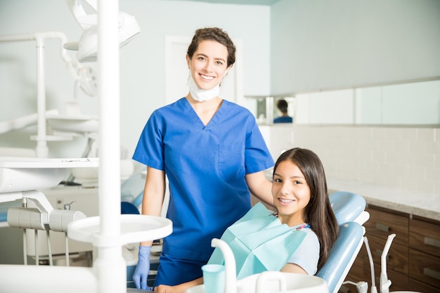 Portret van een glimlachende jonge vrouwelijke tandarts die bij een tienermeisje in de kliniek staat