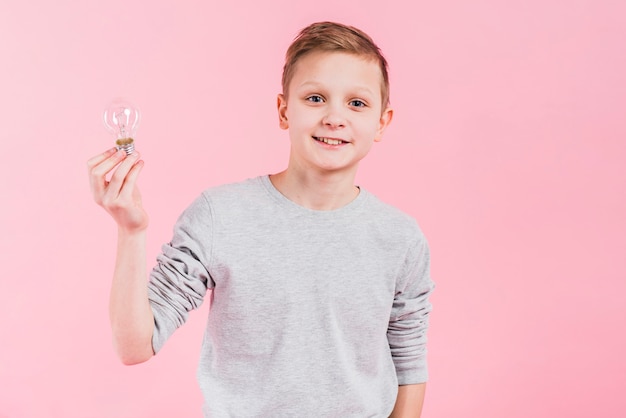 Portret van een glimlachende jongen die gloeilamp houden die in hand zich tegen roze achtergrond bevinden