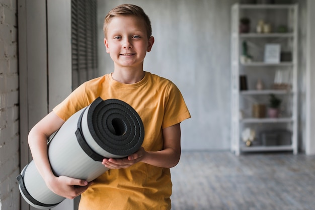 Portret van een glimlachende jongen die grijs opgerolde oefeningsmat ter beschikking houdt