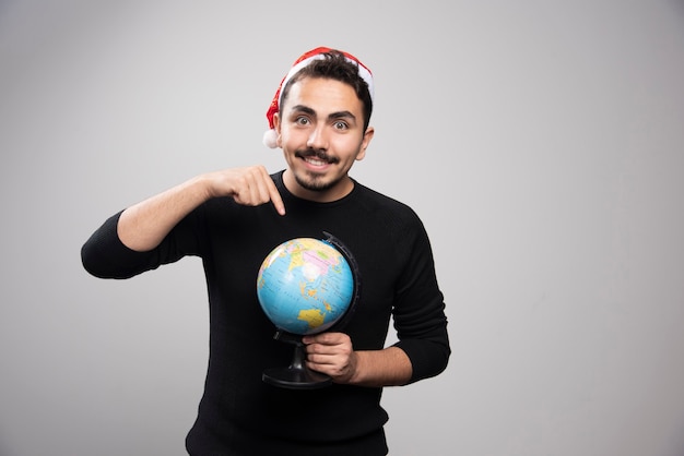 Portret van een glimlachende man in de hoed van de Kerstman wijzend op een wereldbol.