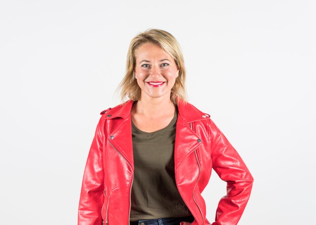 Portret van een glimlachende modieuze blonde rijpe vrouw in rood die jasje op witte achtergrond wordt geïsoleerd