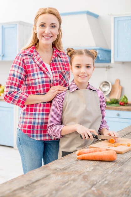 Portret van een glimlachende moeder en een dochter die de wortel met mes in de keuken snijden
