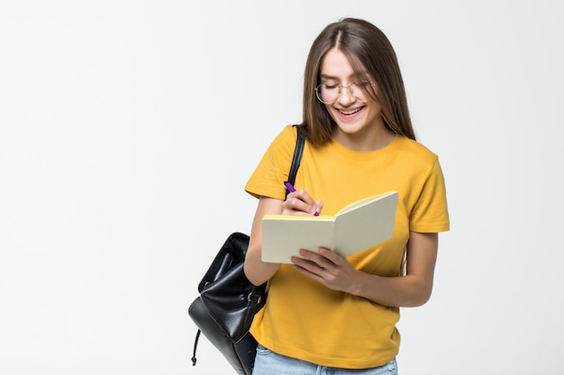 Portret van een glimlachende toevallige studente die met rugzak in een blocnote schrijven terwijl status met boeken over witte muur worden geïsoleerd