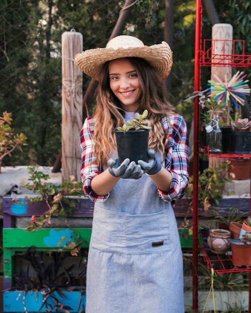 Portret van een glimlachende vrouwelijke tuinman die hoed dragen die cactusinstallatie in de tuin tonen