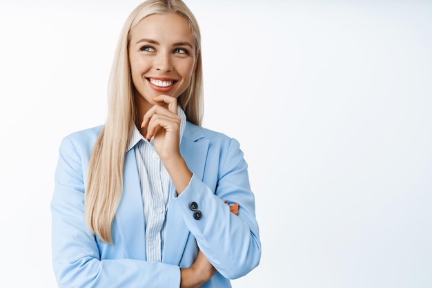 Portret van een glimlachende zelfverzekerde zakenvrouw die denkt en er geïntrigeerd uitziet terwijl ze in een pak staat op een witte achtergrond