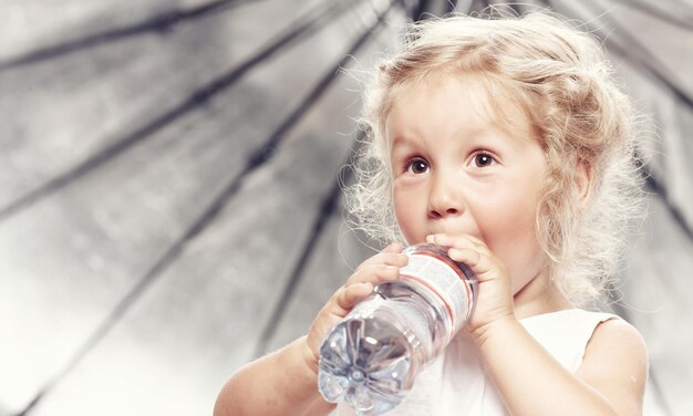 Portret van een grappig schattig klein meisje in een casual jurk, drink water terwijl ze in een studio zit.