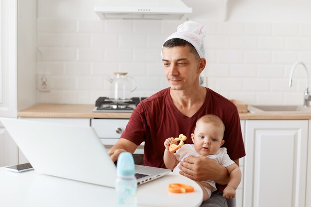 Portret van een grappige, geconcentreerde knappe freelancer met een bordeauxrood t-shirt, poserend in een witte keuken, achter een laptop zittend met een baby in handen en werkend.