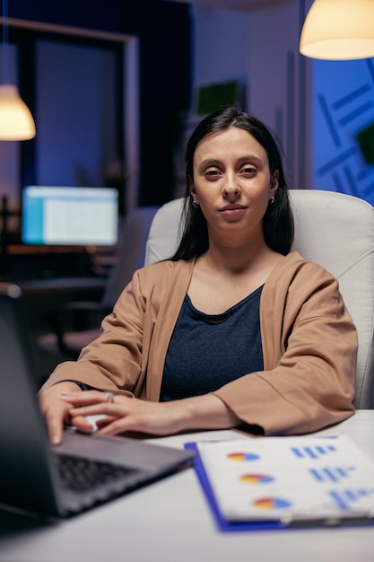 Portret van een hardwerkende manager die naar de camera kijkt en overuren maakt. Slimme vrouw zit op haar werkplek in de loop van de late nachturen en doet haar werk.