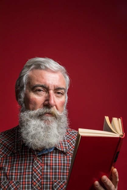 Portret van een hogere mens met grijze baard die aan het boek van de cameraholding in hand tegen rode achtergrond kijken