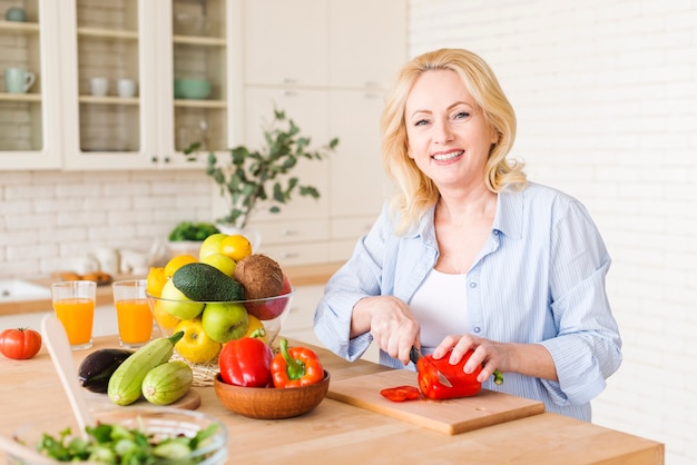 Portret van een hogere vrouw die de rode groene paprika met mes op hakbord in de keuken snijdt