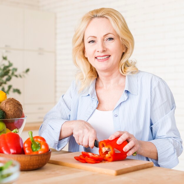 Portret van een hogere vrouw die de rode groene paprika met mes op hakbord snijdt