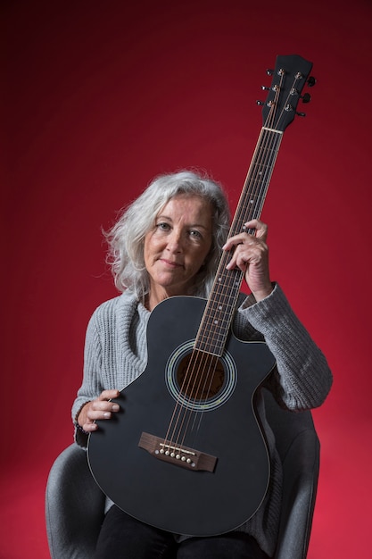 Portret van een hogere vrouwenzitting op de gitaar van de stoelholding tegen rode achtergrond