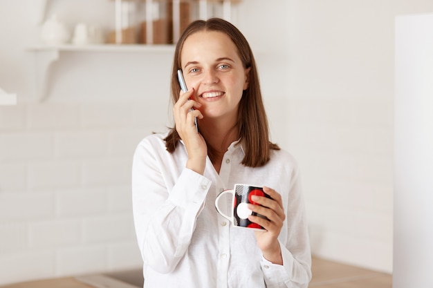 Portret van een innemende vrouw met donker haar die via een smartphone praat, een kopje in handen houdt, geniet van koffie of thee terwijl ze een aangenaam gesprek heeft, naar de camera kijkt.