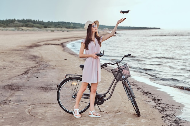 Portret van een jong brunette meisje met een zonnebril en hoed die een jurk draagt, geniet van de vakantie op het strand, spelend met de quadcopter.