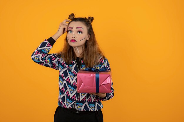 Portret van een jong meisje dat creatieve make-up draagt en een geschenkdoos op een oranje achtergrond houdt. Hoge kwaliteit foto