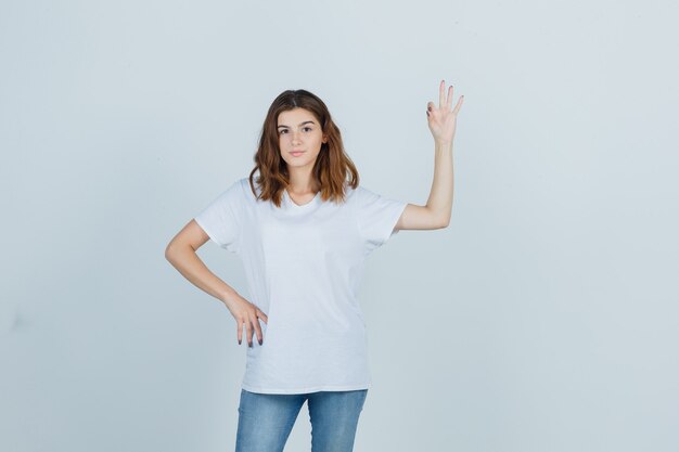 Portret van een jong meisje dat ok gebaar in wit t-shirt, spijkerbroek toont en zelfverzekerd vooraanzicht kijkt