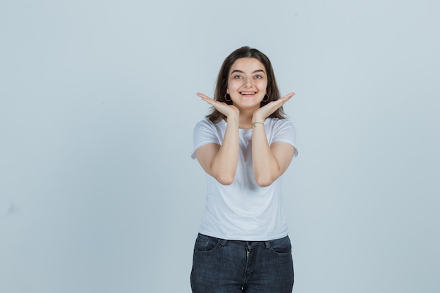 Portret van een jong meisje hand in hand in de buurt van kin in t-shirt, spijkerbroek en op zoek schattig vooraanzicht