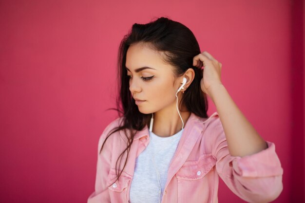 Portret van een jong mooi meisje met donker haar dat staat en naar muziek luistert in oortelefoons terwijl ze bedachtzaam opzij staat op een roze achtergrond