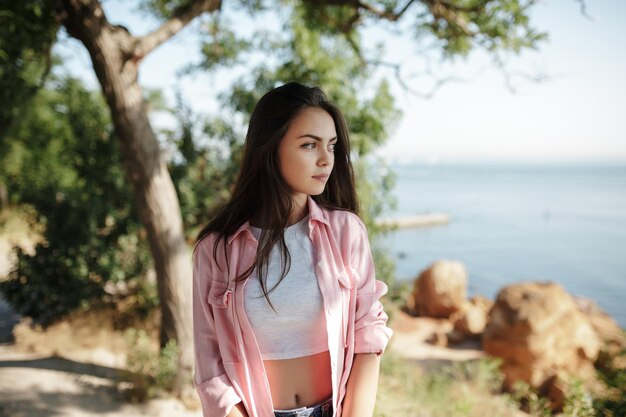 Portret van een jong mooi meisje met donker haar in een roze shirt dat staat en dromerig opzij kijkt met prachtig uitzicht op zee op de achtergrond