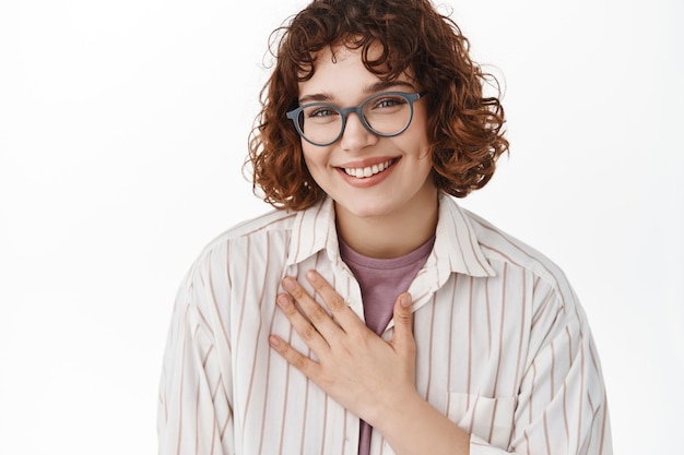 Portret van een jong openhartig meisje met een bril houdt de hand op de borst en glimlacht oprecht naar de camera, lacht en uiting van natuurlijke echte emoties, staande op wit