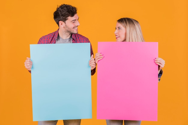 Portret van een jong paar die blauw en roze aanplakbiljet houden tegen een oranje achtergrond