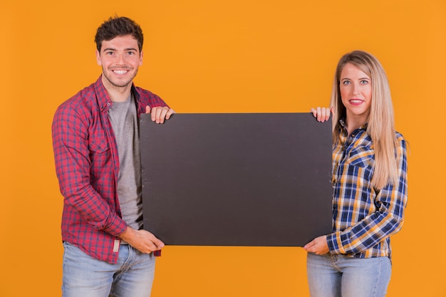 Portret van een jong paar die leeg zwart aanplakbiljet op tegen een oranje achtergrond houden