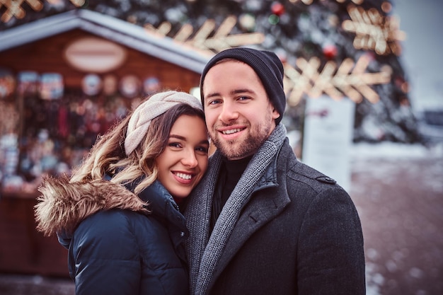 Portret van een jong stel met warme kleren in de buurt van een kerstboom in de stad, genietend van tijd samen doorbrengen, glimlachen en kijken naar een camera. Feestdagen, Kerstmis, Wintertijd.