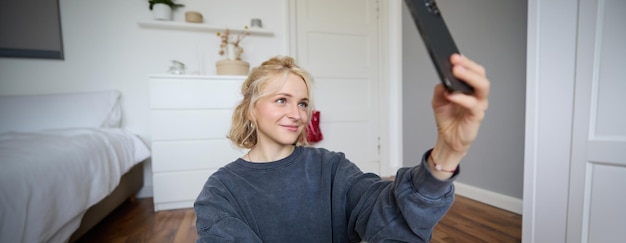 Gratis foto portret van een jong stijlvol meisje dat op de vloer van de slaapkamer zit en selfies maakt met haar smartphone.