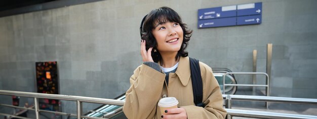 Portret van een jong studentenmeisje in een koptelefoon drinkt koffie en staat op straat met rugzak-pendels