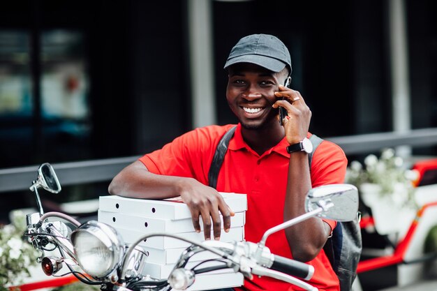 Portret van een jonge afrikaanse man accepteert de bestelling telefonisch in een motorfiets met dozen met pizza en zit op zijn fiets. Stedelijke plek.