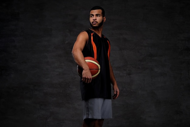 Portret van een jonge Afro-Amerikaanse basketballer in sportkleding geïsoleerd over donkere achtergrond.