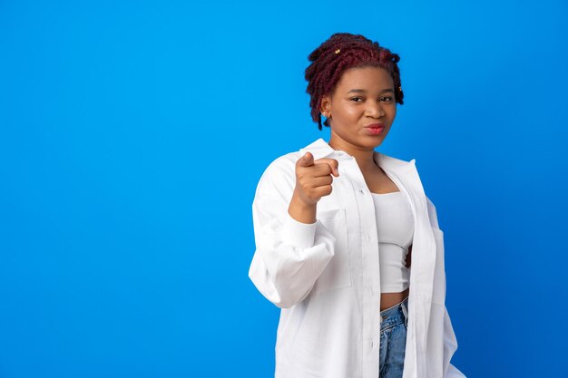Portret van een jonge afro-vrouw die op je wijst tegen een blauwe achtergrond