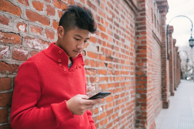 Portret van een jonge Aziatische man met zijn mobiele telefoon buitenshuis tegen bakstenen muur. Communicatie concept.