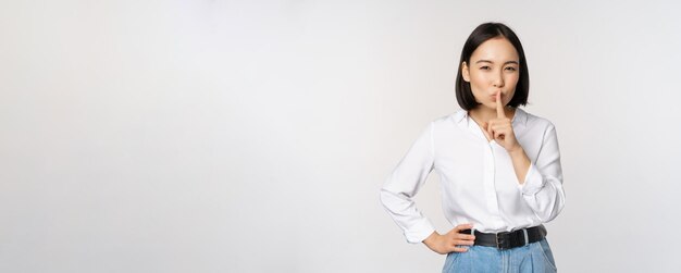 Portret van een jonge aziatische volwassen vrouw die zwijgt, druk op de vinger naar de lippen en deelt geheim, spreek geen taboe-gebaar dat op een witte achtergrond staat
