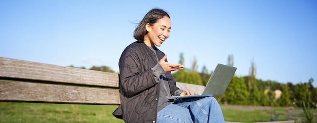 Portret van een jonge Aziatische vrouw die op afstand werkt vanuit het park en op een bankje zit met een laptop die praat