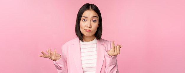 Portret van een jonge Aziatische zakenvrouw die de schouders ophaalt en er verward uitziet, geen idee heeft van iets dat over een roze achtergrond staat