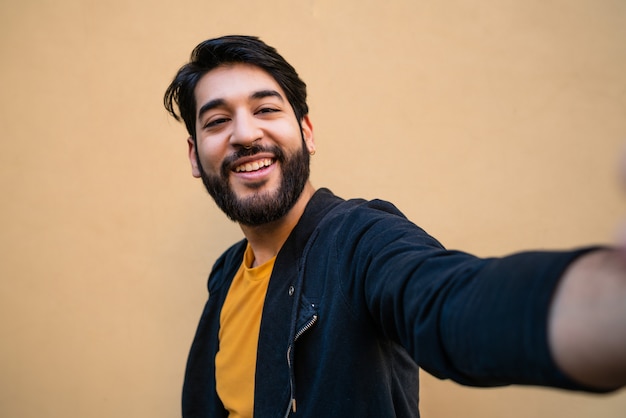 Portret van een jonge bebaarde hipster man camera kijken en het nemen van een selfie tegen geel.