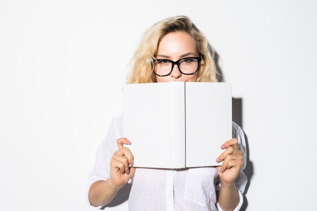 Portret van een jonge bedrijfsvrouw die zich achter een boek met bril verstopt, geïsoleerd op een grijze muur