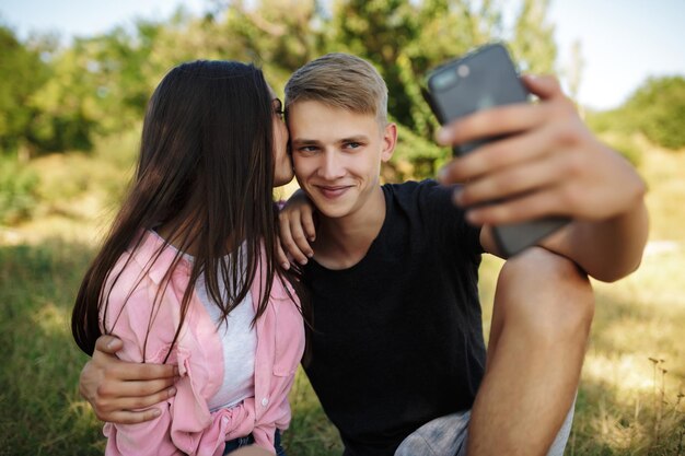 Portret van een jonge coole jongen die op het gazon in het park zit en een meisje omhelst terwijl hij een selfie maakt. Leuk stel dat foto's maakt op de frontale camera van de mobiele telefoon