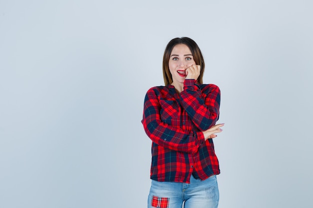 Portret van een jonge dame die de hand op de wang houdt in een geruit overhemd, jeans en een verbaasd vooraanzicht