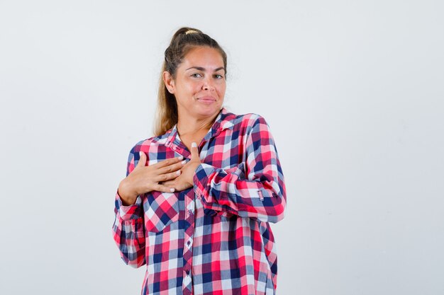 Portret van een jonge dame die de handen op de borst in een geruit overhemd houdt en vrolijk vooraanzicht kijkt