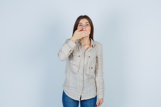 Portret van een jonge dame die de mond bedekt met de hand in casual, jeans en een geschokt vooraanzicht