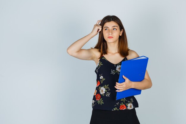 Portret van een jonge dame die een map vasthoudt terwijl ze haar hoofd krabt in blouse, rok en besluiteloos vooraanzicht kijkt