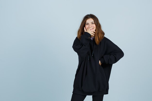 Portret van een jonge dame die hand op mond houdt terwijl hand op taille in oversized hoodie, broek en gelukkig vooraanzicht kijkt