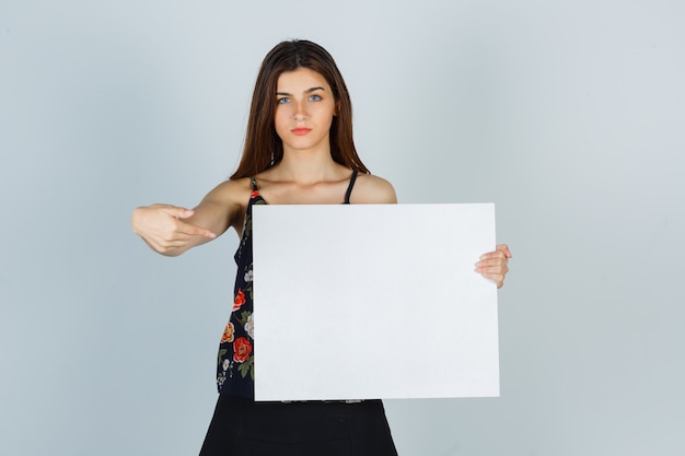 Portret van een jonge dame die naar een leeg canvas in blouse, rok wijst en er serieus vooraanzicht uitziet